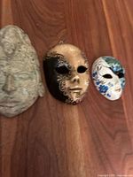 Three ceramic decorative masks laid on a wooden surface: abstract face mask, gold/black Venetian mask, and small floral mask.