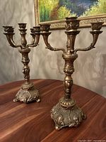 Photo of the pair of brass candelabras on a wooden surface against a beige wall, showing overall form and height.