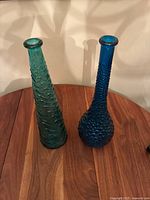 Two tall art glass vases - left teal with swirl texture, right blue with honeycomb texture - on a wooden table.