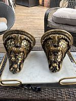 Photo of both gold wooden decorative wall brackets placed side by side on a marble tray, showing detailed face carving and scale pattern with minor scratches.
