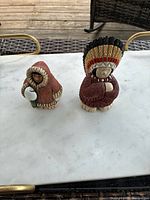 Two ceramic sculptures placed on a white-marble-like surface, one dressed in a hooded garment holding a white object, and the other wearing a feathered headdress.