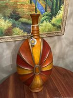 Full view of the large multicolor ceramic vase showing sunburst design and attached ornament, placed in front of a landscape painting.