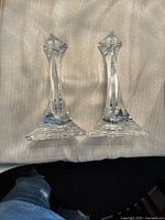 Two clear crystal candleholders shown from above on a light-colored fabric surface. The candleholders feature sleek, faceted designs with square bases and pointed tops.