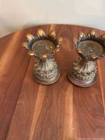 Pair of ornate metal candle holders displayed on wooden surface showing flower petal design on top and a detailed base with leaf motifs.