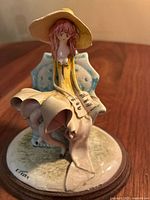 Porcelain figurine showing seated girl in yellow hat and shawl on blue chair with wooden base. The signature 'E. 1979' is visible on the base.