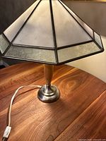 Stained glass lamp shade with geometric white and textured panels and bronze frame sits on a wood-grain table.