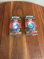 Two unopened TDK QS 8-hour VHS videotapes on wooden surface, front view showing box design and branding.