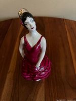Front angled view of vintage 1930s Art Deco porcelain figurine of a woman tightly holding a fan behind her back, wearing a red dress with ruffles, black hair styled up with a pink flower accessory, painted facial features.