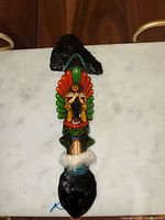 Full view of the vintage handmade Aztec warrior ax on white surface, showing the overall shape and structure with black ends and colorful warrior figure in the middle.