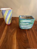 Two handmade vintage vases on a wood surface, one triangular with pastel stripes, the other square with green textured glaze.
