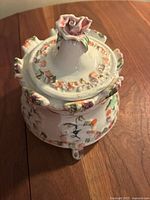 Front angled view of the vintage porcelain container showing floral relief details and rose handle on lid.