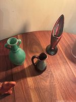 Photo showing three vintage pottery items on wood surface: green vase with handles, small jug, tall red narrow vase