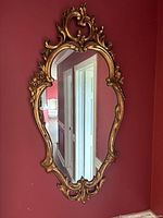 Baroque style ornate wall mirror with gilded gold carved wooden frame hanging on a red wall. Shows detailed raised floral and scroll motifs, elongated oval shape with ornamental crest on top.