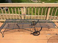 Photo of black wrought iron chaise lounge with mesh design positioned on wooden deck, showing its full length and curved shape with wheels and handle.
