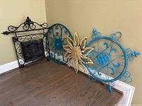 Four pieces of decorative metal wall art with sun motif and geometric designs in black, blue, and gold colors shown leaning against a wall on hardwood floor.