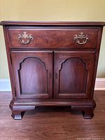 Front view of wooden nightstand with one drawer and two cabinet doors. Brass handles on drawer and cabinets visible, showing shape and detailing.