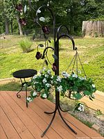 Black decorative iron plant stand with metal flower decorations and hanging basket containing trailing white flowers positioned beside round black metal table on outdoor deck.