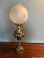 Full view of the ornate brass lamp with frosted glass globe on a plain background, showing front and base detail.