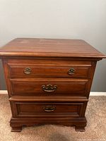 Front view of the wooden nightstand showing three drawers with brass handles, carved feet, and wood finish.