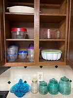 Open kitchen wooden cabinet containing stacked white ceramic dishes, several plastic storage containers with lids in red, purple, blue, and clear. Four vintage textured blue-green glass canisters with lids and one clear glass jar without lid on counter. Small blue carnival glass basket on counter.