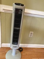 Full view of gray Lasko tower fan showing vertical air vents and fan base with power cord coiled beside it.