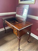 Front and angled side view of wooden vanity table with flip-top mirror open showing storage compartments, Queen Anne style cabriole legs, and brass hardware.