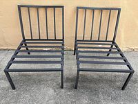 Pair of modern style black metal outdoor chairs with horizontal slat seating and vertical slat backs, shown from front angle on concrete surface against a yellowish wall.