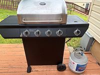 Full frontal view of Nexgrill propane grill on deck with propane tank beside it showing all 5 control knobs and lower stand with wheels.