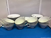 Eight coffee cups arranged partly stacked with eight saucers and other plates in the background, showing white china with floral design in silver trim.
