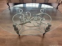 Top-down view showing clear oval glass top and intricate metal scroll base design with floral accents.