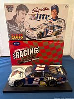 Front angled photo of diecast model car, box featuring Rusty Wallace and Elvis Presley images and signatures, displayed on blue fabric surface.