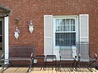Full set of black metal outdoor patio furniture arranged against brick wall: glider bench, two chairs, two tables.