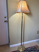 Full view of the traditional brass floor lamp with beige fabric shade lit up, showing height and style against wall and door.