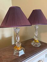 Pair of traditional glass table lamps with deep purple, pleated shades placed on a wooden surface, showing clear detailed base and overall condition.