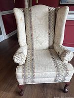 Front view of traditional wingback chair featuring off-white damask patterned fabric and padded armrests.