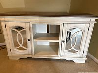 Front view of the wooden cabinet showing white distressed finish with brown trim, decorative mirrored doors with wooden overlays and black knobs, open center shelves.