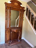 Front angle view of wooden traditional hall tree against wall, showing mirror, decorative top, shelf, and hooks.