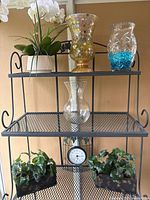 Black metal shelf displaying faux white orchid plant, decorative mosaic glass vase, clear vase with blue beads, clear hurricane vase, analog clock, and two rectangular planters with faux greenery.