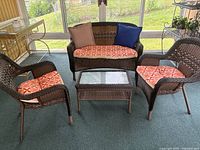 Front view of two brown plastic wicker chairs with orange patterned cushions, loveseat with cushions and pillows, and rectangular glass-top wicker table indoors near sunroom windows.