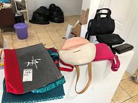 Assorted tote bags, handbags, and purses displayed on a white surface including a grey felt tote bag with red accents, cream and blush handbag, black duffel bag, and several folded wallets.