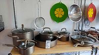 Full set on wooden shelf showing metal cookware with glass lids and one frying pan. Green silicone lids and metal strainer also hung above.