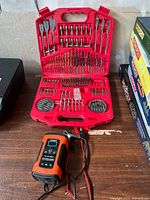 Red plastic case open with assorted drill bits, spade bits, and driver bits neatly organized inside. Foxsur battery charger placed in front of case on wooden surface.