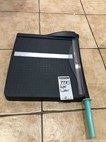 Full view of a black guillotine paper cutter on a tiled floor, showcasing the turquoise cutting handle and metal ruler on the base.