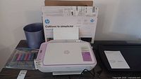 Front view of the HP DeskJet 2842e printer sitting on a blue lap desk. Original HP box, ink cartridges, manuals, and a purple wastebasket are also visible.