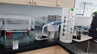 Wide view of kitchen corner showing white plastic tiered dish rack, glass baking dish, Brita pitcher, white bottle holder, and parts of other items.
