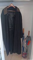 Men's black Carrington coat hanging on a hanger, showing full length and inner lining. Next to it is an umbrella holder containing multiple umbrellas.
