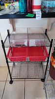 Front view of shelving unit showing two clear plastic baskets on second shelf and two red plastic baskets on third shelf, black metal frame with wire shelves and caster wheels.