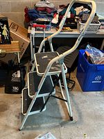 Side angle view of the metal utility step ladder fully opened showing the curved handle and three steps with black treads.