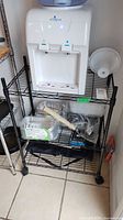 Front view of metal wire shelving unit with a water dispenser on top shelf and miscellaneous items on other shelves.