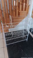 Photo showing front and side view of white metal wire shoe rack standing on tile floor next to wooden stairs and black mat.